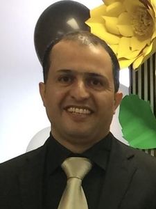 A man wearing a black suit and gold tie smiles in front of a wall decorated with large paper flowers and balloons.