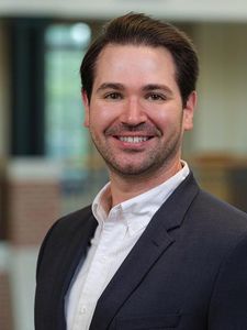 A man with dark hair and light facial hair smiles while wearing a dark blazer and white shirt against a blurred indoor.