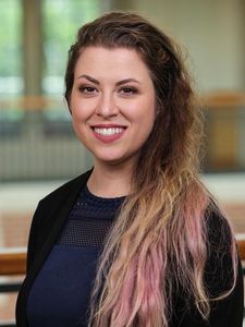 A woman with long wavy hair dyed blonde and pink smiles while wearing a black cardigan and navy top indoors.