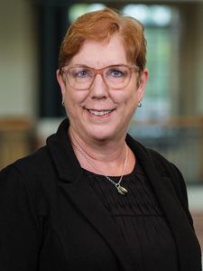 A person with short red hair and glasses smiles while wearing a black jacket and necklace against a blurred background.