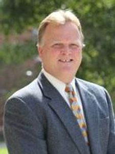 A man with light hair wearing a gray suit and patterned tie stands outdoors with greenery blurred in the background.
