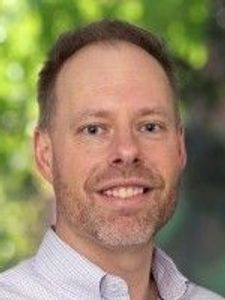 A man with short light brown hair and a trimmed beard smiles wearing a light purple checkered shirt against a blurred green.