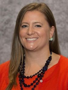A woman with long light brown hair smiles at the camera wearing a red top and a layered dark blue beaded necklace.
