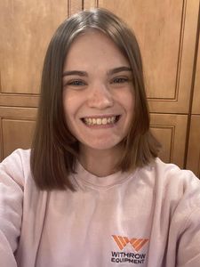 A young person with shoulder-length brown hair smiles at the camera wearing a light pink sweatshirt with orange and black.