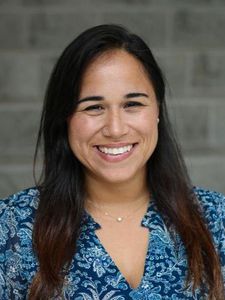 A woman with long dark hair smiles at the camera wearing a blue patterned blouse against a gray brick background.