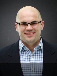 A bald man wearing glasses, a checkered shirt, and a dark blazer smiles against a plain dark background.