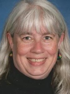 A smiling woman with long gray hair and bangs wears colorful earrings and a black top against a blue background.