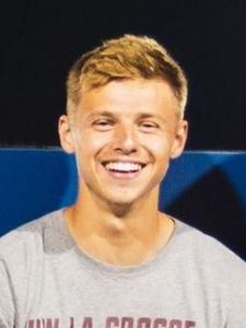 A young man with short blond hair is smiling and wearing a light gray shirt against a dark background.