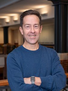 A man in a blue sweater smiles with his arms crossed, standing indoors near a railing with blurred background lights.