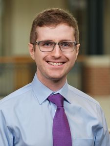 A man with short brown hair and glasses smiles while wearing a light blue shirt and purple tie in an indoor setting.