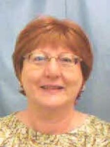 A middle-aged woman with short reddish hair and glasses smiles gently against a plain light blue background.