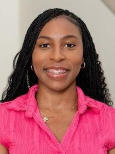 A woman with long braided hair smiles wearing braces, gold hoop earrings, a gold necklace, and a bright pink blouse.