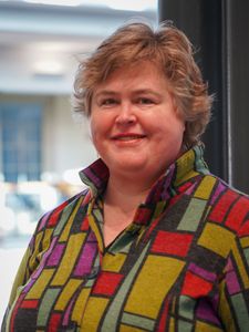 A person with short wavy hair smiles while wearing a colorful geometric patterned jacket indoors.