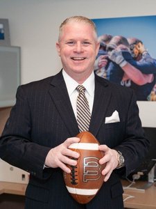 A smiling JCU faculty member in a suit holds a football. In the background, students celebrate a sports victory.