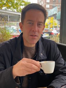 A man wearing a dark jacket holds a small white cup while sitting at an outdoor café table on a city street.