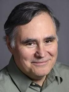 A middle-aged man with dark hair and gray sideburns smiles gently while wearing a green collared shirt against a plain gray.