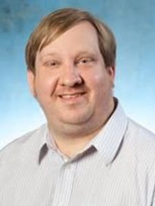 A man with light brown hair and fair skin smiles gently while wearing a light striped shirt against a blue background.