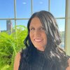A JCU faculty member with long dark hair smiles warmly for a portrait, standing indoors with a campus view outside a window.