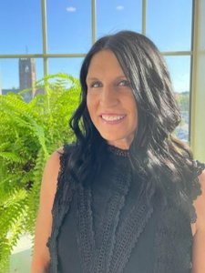 A JCU faculty member with long dark hair smiles warmly for a portrait, standing indoors with a campus view outside a window.