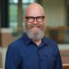 A smiling bald man with a full gray beard and glasses wearing a blue button-up shirt stands indoors.