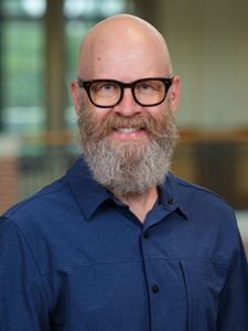 A smiling bald man with a full gray beard and glasses wearing a blue button-up shirt stands indoors.