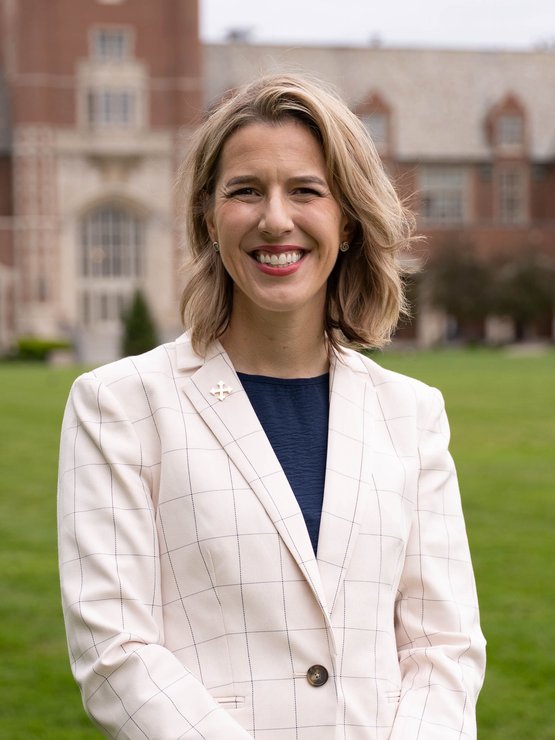 A smiling woman with shoulder-length blonde hair wears a white checked blazer and stands on a green lawn with a brick.