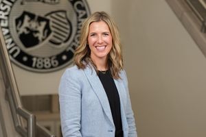 A smiling woman with shoulder-length blonde hair wears a light blue blazer and stands indoors by a staircase.