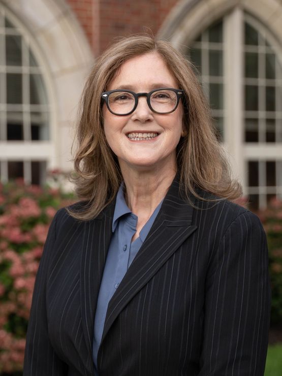 A portrait of a woman with blonde/light brown hair, wearing glasses, a blue shirt, and a dark pinstriped blazer.