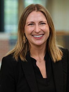 A woman with shoulder-length blonde hair smiles at the camera wearing a black blazer and earrings.