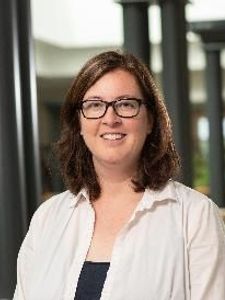 A woman with shoulder-length brown hair and glasses smiles while standing indoors near black columns and a brick wall.