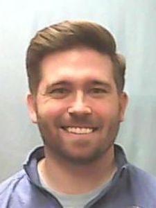 A man with short brown hair and a beard smiles while wearing a blue jacket against a plain light background.
