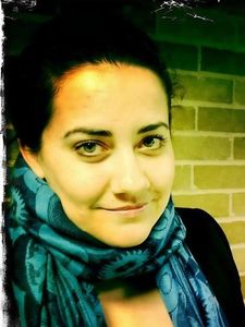 A woman with dark hair pulled back wears a blue patterned scarf and a black top, smiling slightly against a brick wall.