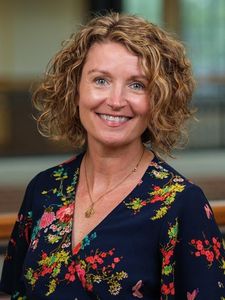 A woman with curly blonde hair smiles at the camera wearing a dark floral blouse and a gold necklace.