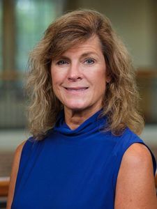 A woman with shoulder-length wavy light brown hair wearing a sleeveless blue top smiles softly against a blurred indoor.