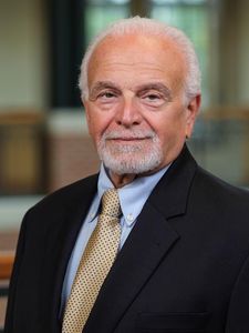 An older man with white hair and a trimmed beard wears a black suit jacket, light blue shirt, and patterned tie.