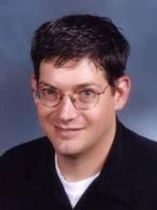 A man with short dark hair and glasses smiles slightly while wearing a black shirt against a blue-gray background.