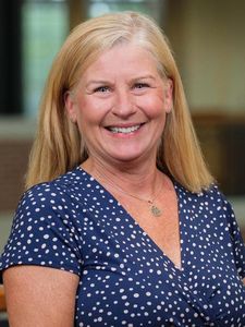 A smiling woman with straight blonde hair wears a blue dress with white polka dots and a round pendant necklace.