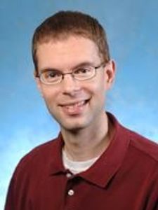 A man with short brown hair and glasses smiles while wearing a maroon polo shirt against a blue background.