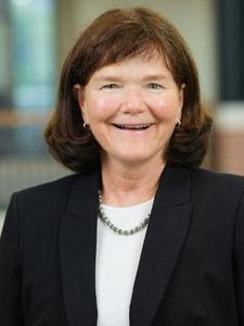 A woman with short brown hair smiles while wearing a black blazer, light pink top, and beaded necklace indoors.