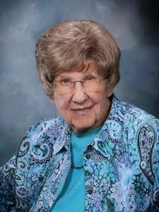 An elderly woman with short light brown hair and glasses smiles gently while wearing a blue patterned jacket over a teal top.