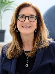 A smiling woman with shoulder-length light brown hair wears black glasses, silver earrings, a necklace, and a dark blazer.