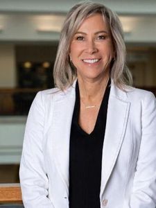A woman with shoulder-length blonde hair and glasses smiles while wearing a floral blouse and dark cardigan.