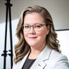 A woman with wavy light brown hair and glasses smiles slightly while wearing a light gray blazer and black top.