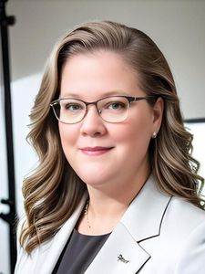 A woman with wavy light brown hair and glasses smiles slightly while wearing a light gray blazer and black top.