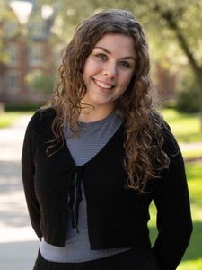 A woman with long curly hair smiles warmly while standing outdoors on a sunny day with blurred trees and buildings behind.