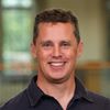 A man with short brown hair smiles at the camera wearing a dark collared shirt with a blurred indoor background.