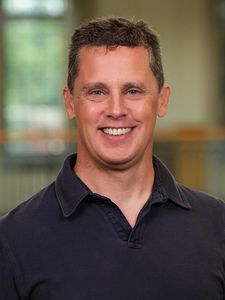 A man with short brown hair smiles at the camera wearing a dark collared shirt with a blurred indoor background.