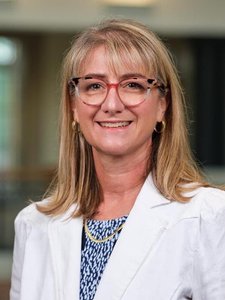 A woman with shoulder-length blonde hair and glasses smiles while wearing a white jacket and patterned top indoors.