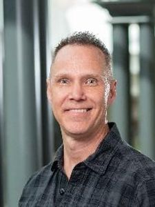 A man with short curly hair and a dark checkered shirt smiles while standing in a corridor with columns and blurred.