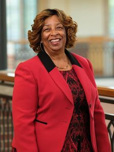 A smiling woman with curled hair wears a red blazer over a patterned dress standing indoors near a railing.